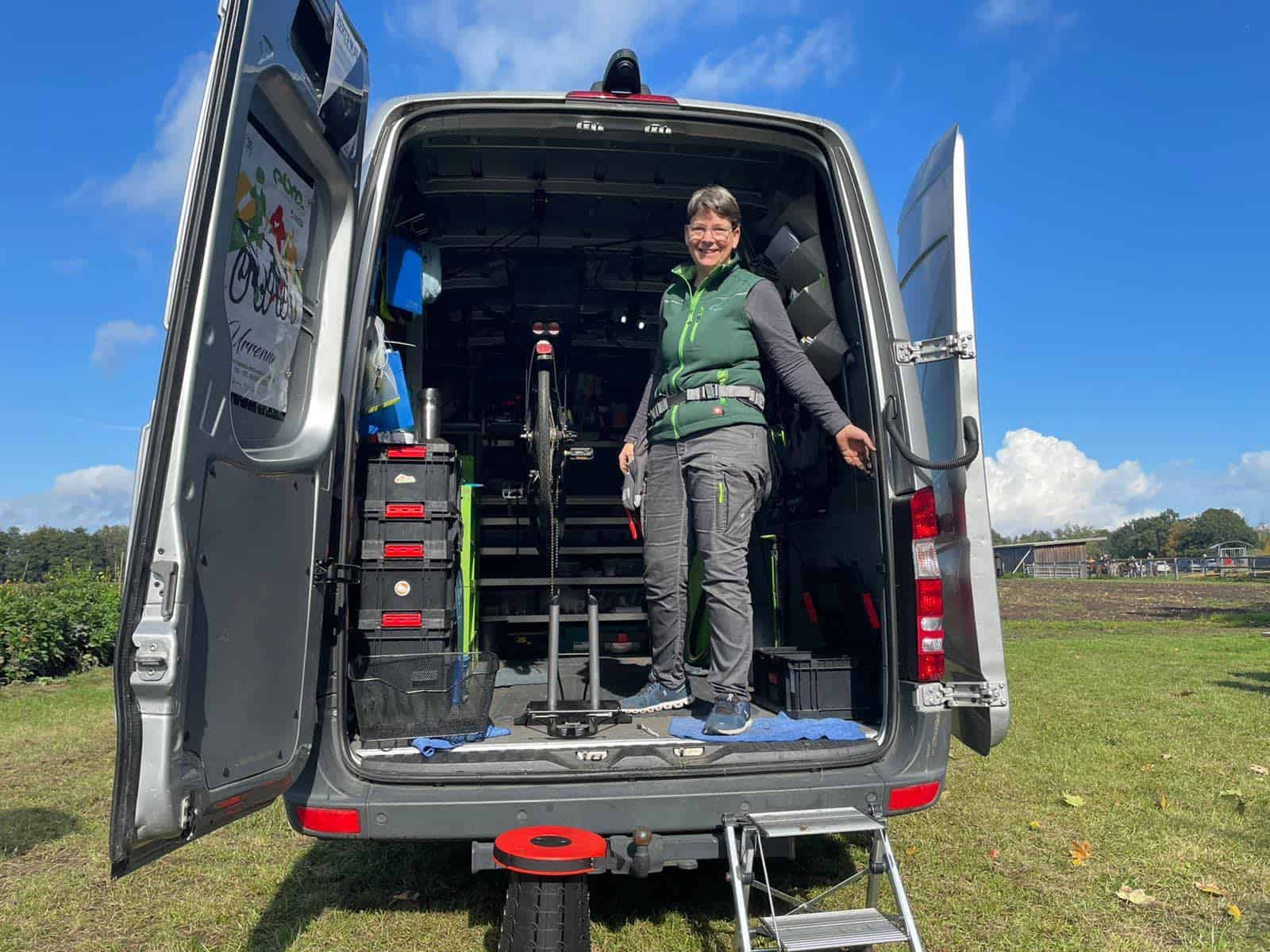 Es ist das innere eines Sprinters zu sehen. Kisten und Regale mit Werkzeug und Fahrradteilen sind zu sehen. Michaela Mocke steht in einer schwarzen Hose, grauem Langarmshirt und grüner Weste lächelnd auf der Ladefläche.